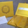 A high-angle shot of a luxurious wedding invitation suite in vibrant gold, arranged on a soft white fabric. * **On the left:** An open gold silk invitation box. The inner lid features a pocket holding a white RSVP card with gold floral accents. The bottom compartment contains a clear acrylic invitation card. * **On the right:** The closed lid of the gold box, featuring an embossed circular emblem with ornate scrollwork, the names "James Parker & Karen Brett," the date "09.09.2019," and small embedded crystals. * **In the foreground:** A white mailing envelope addressed to "Aleeya Rahman" in gold lettering. * **In the background:** Decorative gold lace pieces with small blue crystals are scattered for additional texture.
