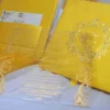 A high-angle, close-up shot of a luxury wedding invitation suite in shimmering gold. A clear acrylic invitation card with gold-foil script and ornate corner flourishes for "Patrick and Prisca" is leaned against its matching gold silk box. The lid of the box, visible behind the acrylic, features a debossed circular crest decorated with crystals. To the left, the open base of the box shows a yellow-gold satin lining and an RSVP card tucked into an interior pocket. The entire suite rests on a soft, white textured fabric.
