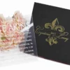 An overhead, angled shot of a luxury wedding invitation suite. To the left is a closed, square black box featuring a metallic gold-foil fleur-de-lis emblem and the names "Cyntoria and Leroy" in elegant script. To the right, an open version of the same box reveals a vibrant red suede-like interior lining holding a clear acrylic invitation card with white printed text. A bouquet of soft pink roses in the top right corner adds a romantic touch against the clean white background.