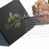 An overhead, angled shot of a luxury wedding invitation suite. To the left is a closed, square black box featuring a metallic gold-foil fleur-de-lis emblem and the names "Cyntoria and Leroy" in elegant script. To the right, an open version of the same box reveals a vibrant red suede-like interior lining holding a clear acrylic invitation card with white printed text. A bouquet of soft pink roses in the top right corner adds a romantic touch against the clean white background.