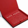 An open, luxurious invitation box with a black exterior and a vibrant red suede-like interior lining. Resting inside the base of the box is a clear acrylic invitation card. The card features bold white and silver text announcing "KATE AKINS" being called to the Torah as a Bat Mitzvah on Friday, April 1, 2022. The text provides event details including the location at Montgomery Ave, Wynnewood, PA, and RSVP information. The image is captured at an angle against a white background.