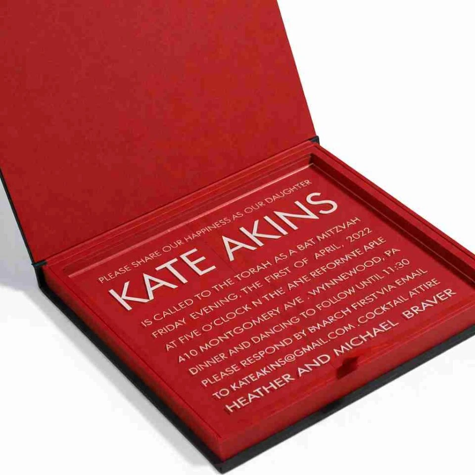 An open, luxurious invitation box with a black exterior and a vibrant red suede-like interior lining. Resting inside the base of the box is a clear acrylic invitation card. The card features bold white and silver text announcing "KATE AKINS" being called to the Torah as a Bat Mitzvah on Friday, April 1, 2022. The text provides event details including the location at Montgomery Ave, Wynnewood, PA, and RSVP information. The image is captured at an angle against a white background.