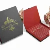 An angled, close-up shot of a closed, square black invitation box featuring a metallic gold-foil fleur-de-lis emblem in the center. Overlapping the emblem is the elegant gold-script text "Cyntoria and Leroy" along with the wedding date "8.18.18." A hint of a red interior lining is visible along the side edge. In the upper right corner, a soft bouquet of light pink roses serves as a decorative background element against the white surface.