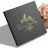An angled, close-up shot of a closed, square black invitation box featuring a metallic gold-foil fleur-de-lis emblem in the center. Overlapping the emblem is the elegant gold-script text "Cyntoria and Leroy" along with the wedding date "8.18.18." A hint of a red interior lining is visible along the side edge. In the upper right corner, a soft bouquet of light pink roses serves as a decorative background element against the white surface.