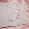 A high-angle shot of a refined Bat Mitzvah invitation for partially inserted into a matching white envelope. The invitation features a rose gold metallic font and a **Star of David** symbol, bordered by intricate floral swirls and a delicate dotted line. The set is displayed on a backdrop of luxuriously draped **blush pink satin** fabric, highlighting the card's elegant and feminine aesthetic.