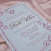 An elegant Bat Mitzvah invitation for featuring a clean white card with rose gold or copper metallic text. The design includes at the top and is framed by delicate, swirling floral illustrations and a dotted border in a matching metallic finish. The invitation is captured at an angle, resting on a soft, draped **mauve-pink satin** fabric.