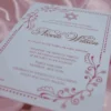 An elegant Bat Mitzvah invitation for featuring a clean white card with rose gold or copper metallic text. The design includes at the top and is framed by delicate, swirling floral illustrations and a dotted border in a matching metallic finish. The invitation is captured at an angle, resting on a soft, draped **mauve-pink satin** fabric.