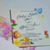 An elegant White Acrylic Wedding Invitations Floral Artwork with envelope is shown in a close-up, angled shot, leaning against a lush bouquet of soft peach and pink roses. The invitation features an intricate white laser-cut floral border that surrounds the reflective center. The text is printed in crisp Black.