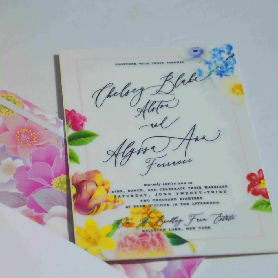 An elegant White Acrylic Wedding Invitations Floral Artwork with envelope is shown in a close-up, angled shot, leaning against a lush bouquet of soft peach and pink roses. The invitation features an intricate white laser-cut floral border that surrounds the reflective center. The text is printed in crisp Black.
