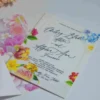 An elegant White Acrylic Wedding Invitations Floral Artwork with envelope is shown in a close-up, angled shot, leaning against a lush bouquet of soft peach and pink roses. The invitation features an intricate white laser-cut floral border that surrounds the reflective center. The text is printed in crisp Black.