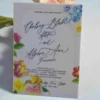 An elegant White Acrylic Wedding Invitations Floral Artwork with envelope stands upright against a bouquet of peach-colored roses on a white surface. The invitation features a vibrant, multi-colored floral design with illustrations of pink roses, yellow tulips, and blue wildflowers surrounding the edges