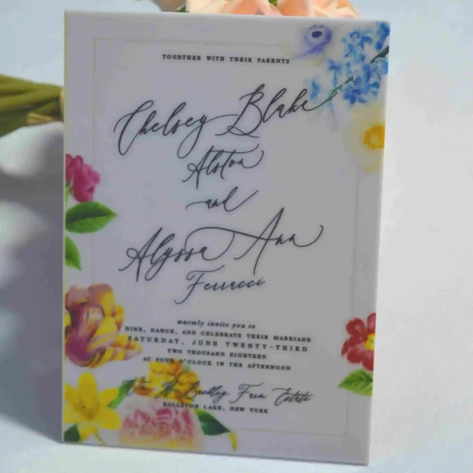 An elegant White Acrylic Wedding Invitations Floral Artwork with envelope stands upright against a bouquet of peach-colored roses on a white surface. The invitation features a vibrant, multi-colored floral design with illustrations of pink roses, yellow tulips, and blue wildflowers surrounding the edges