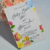 An elegant White Acrylic Wedding Invitations Floral Artwork with envelope stands upright against a bouquet of peach-colored roses on a white surface. The invitation features a vibrant, multi-colored floral design with illustrations of pink roses, yellow tulips, and blue wildflowers surrounding the edges