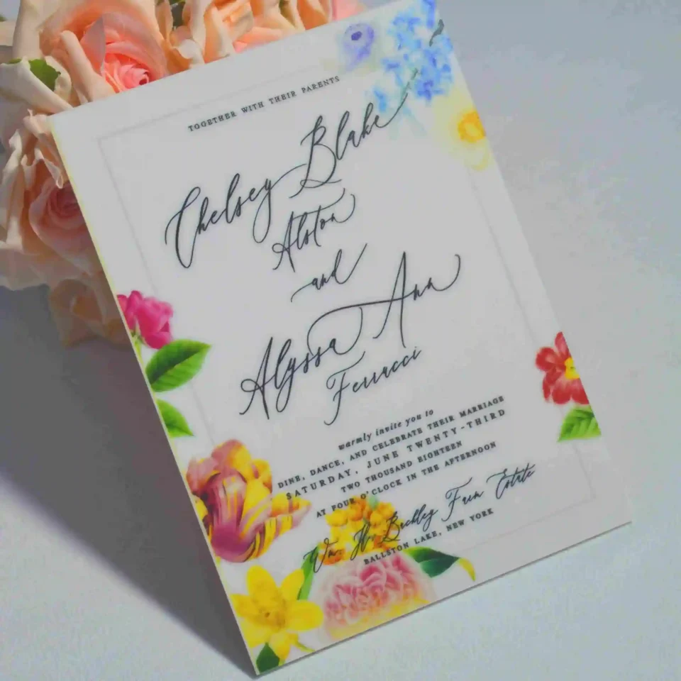 An elegant White Acrylic Wedding Invitations Floral Artwork with envelope stands upright against a bouquet of peach-colored roses on a white surface. The invitation features a vibrant, multi-colored floral design with illustrations of pink roses, yellow tulips, and blue wildflowers surrounding the edges
