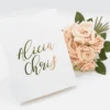 An overhead shot of a wedding invitation suite on a white surface. To the left is a matte white invitation box featuring a gold-foil leaf wreath and an "A" monogram on the lid, accented with gold metal corner caps. To the right, a clear acrylic invitation card rests against a bouquet of soft pink and peach roses. The acrylic card is engraved with golden text for the wedding of featuring delicate floral corner flourishes.