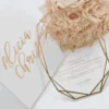 An overhead, vertical shot of a square, matte white invitation box on a white surface. The lid features a centered, metallic gold-foil monogram of the letter "A," surrounded by a circular frame of delicate gold-foil leaves. In the top right corner, a bouquet of soft pink and peach roses adds a gentle floral accent and polished gold metal caps protecting the bottom corners.