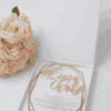 An overhead, vertical shot of a square, matte white invitation box on a white surface. The lid features a centered, metallic gold-foil monogram of the letter "A," surrounded by a circular frame of delicate gold-foil leaves. In the top right corner, a bouquet of soft pink and peach roses adds a gentle floral accent and polished gold metal caps protecting the bottom corners.