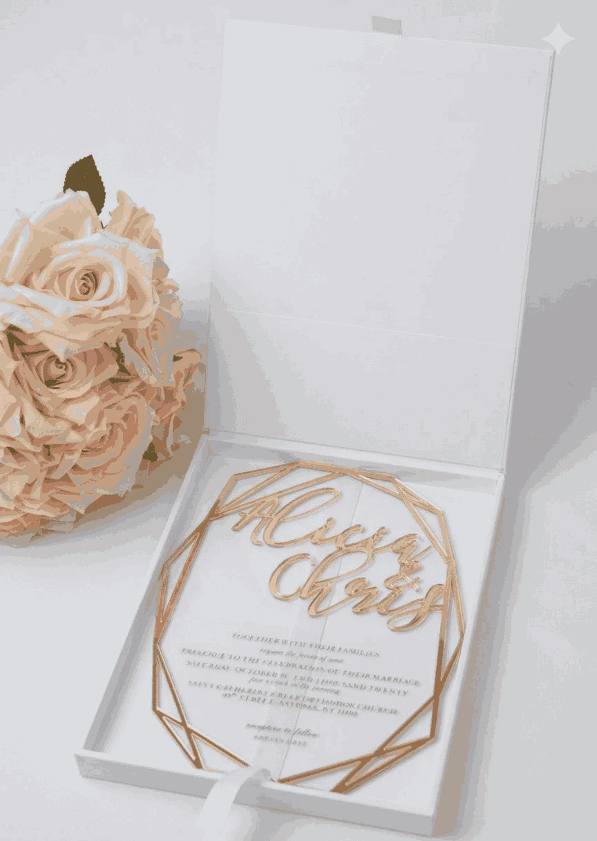 An overhead, vertical shot of a square, matte white invitation box on a white surface. The lid features a centered, metallic gold-foil monogram of the letter "A," surrounded by a circular frame of delicate gold-foil leaves. In the top right corner, a bouquet of soft pink and peach roses adds a gentle floral accent and polished gold metal caps protecting the bottom corners.