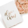 An overhead shot of a wedding invitation suite on a white surface. To the left is a matte white invitation box featuring a gold-foil leaf wreath and an "A" monogram on the lid, accented with gold metal corner caps. To the right, a clear acrylic invitation card rests against a bouquet of soft pink and peach roses. The acrylic card is engraved with golden text for the wedding of featuring delicate floral corner flourishes.