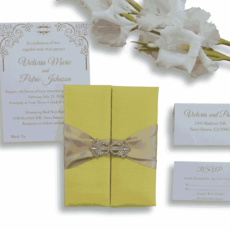 A rectangular, olive-green silk wedding invitation folio oriented vertically. The design features a gate-fold opening with two panels meeting in the center. A shimmering taupe or champagne-colored satin ribbon wraps horizontally around the folio, cinched at the middle by a large, sparkling silver rhinestone brooch with a geometric, butterfly-inspired silhouette. The folio is set against a plain white background, casting a soft shadow at the base.