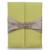 A rectangular, olive-green silk wedding invitation folio oriented vertically. The design features a gate-fold opening with two panels meeting in the center. A shimmering taupe or champagne-colored satin ribbon wraps horizontally around the folio, cinched at the middle by a large, sparkling silver rhinestone brooch with a geometric, butterfly-inspired silhouette. The folio is set against a plain white background, casting a soft shadow at the base.