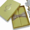 A rectangular, olive-green silk wedding invitation folio oriented vertically. The design features a gate-fold opening with two panels meeting in the center. A shimmering taupe or champagne-colored satin ribbon wraps horizontally around the folio, cinched at the middle by a large, sparkling silver rhinestone brooch with a geometric, butterfly-inspired silhouette. The folio is set against a plain white background, casting a soft shadow at the base.