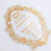 A oval-shaped transparent acrylic wedding invitation with a highly decorative, laser-cut edge. The card is framed by an intricate gold foil baroque border featuring ornate scrolls and flourishes.