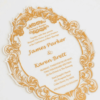 A oval-shaped transparent acrylic wedding invitation with a highly decorative, laser-cut edge. The card is framed by an intricate gold foil baroque border featuring ornate scrolls and flourishes.