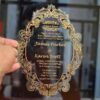 A hand holds an oval-shaped transparent acrylic wedding invitation with a highly decorative, laser-cut edge. The card is framed by an intricate gold foil baroque border featuring ornate scrolls and flourishes.