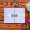 A pale blush pink gatefold wedding invitation resting on a warm, horizontal wood-grain surface. The folder is made of a textured silk or high-quality cardstock and is held closed by a white satin ribbon belly band. At the center of the ribbon is a sparkling silver rhinestone brooch in a butterfly or floral-inspired shape, adding a touch of glamour. To the right, the edge of a white and pink flower is visible, softening the overall composition.