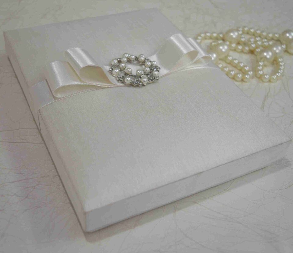 Ivory silk wedding invitation box with a luxury pearl brooch, satin ribbon, and handmade artisan padding.