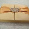 A rectangular, gold silk-textured gatefold invitation box shown from a perspective angle. A wide, shimmering orange satin ribbon wraps horizontally around the box, gathered at the center by an ornate, oval-shaped silver brooch encrusted with clear, sparkling crystals. The box rests on a neutral-toned, textured burlap or linen surface. The lighting emphasizes the fine, horizontal weave of the silk and the warm, metallic tones of the invitation suite.