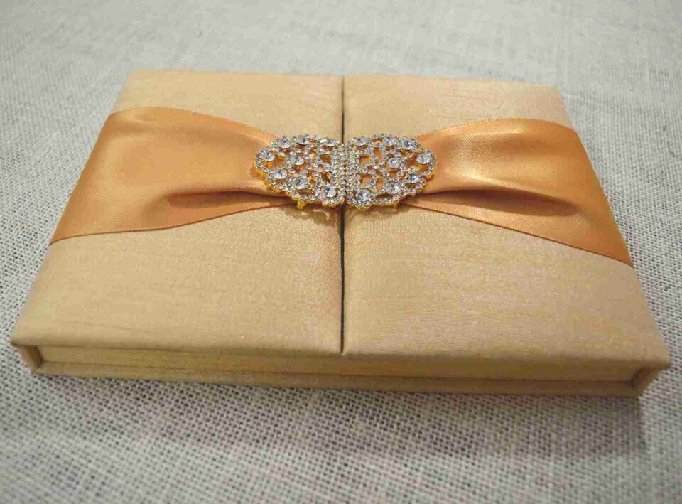 A rectangular, gold silk-textured gatefold invitation box shown from a perspective angle. A wide, shimmering orange satin ribbon wraps horizontally around the box, gathered at the center by an ornate, oval-shaped silver brooch encrusted with clear, sparkling crystals. The box rests on a neutral-toned, textured burlap or linen surface. The lighting emphasizes the fine, horizontal weave of the silk and the warm, metallic tones of the invitation suite.