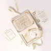 An open, luxurious wedding invitation suite displayed against a white background with flowing ivory ribbons. The main cream-colored box is open, revealing an intricate gold-etched illustration of a Venetian-style palace on the inside lid. Within the box's recessed compartment lies a clear acrylic invitation with gold lettering and a decorative white tassel. Scattered around the box are matching stationary pieces, including a gold-bordered RSVP card and a "Special Note" card, all featuring elegant gold foil flourishes. The closed lid of the box, visible to the left, displays the names under a gold crown crest.