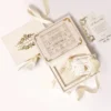 An open, luxurious wedding invitation suite displayed against a white background with flowing ivory ribbons. The main cream-colored box is open, revealing an intricate gold-etched illustration of a Venetian-style palace on the inside lid. Within the box's recessed compartment lies a clear acrylic invitation with gold lettering and a decorative white tassel. Scattered around the box are matching stationary pieces, including a gold-bordered RSVP card and a "Special Note" card, all featuring elegant gold foil flourishes. The closed lid of the box, visible to the left, displays the names under a gold crown crest.An open, luxurious wedding invitation suite displayed against a white background with flowing ivory ribbons. The main cream-colored box is open, revealing an intricate gold-etched illustration of a Venetian-style palace on the inside lid. Within the box's recessed compartment lies a clear acrylic invitation with gold lettering and a decorative white tassel. Scattered around the box are matching stationary pieces, including a gold-bordered RSVP card and a "Special Note" card, all featuring elegant gold foil flourishes. The closed lid of the box, visible to the left, displays the names under a gold crown crest.