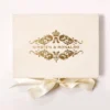 A top-down view of a rectangular, ivory-colored luxury invitation box made of a velvet-like material. The center of the lid features a grand, gold foil crest with intricate floral flourishes and a royal crown at the top. The name printed in a clean, gold serif font in the middle of the design. A wide, ivory satin ribbon is tied in a bow underneath the box, with its ends fanning out against the clean white background.