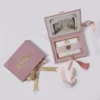 A high-angle view of an open dusty rose velvet wedding invitation box and its coordinating stationery. The inside lid of the box displays a vintage-style illustration of a grand estate framed with gold flourishes. The velvet-lined interior has a recessed compartment and a pink satin ribbon with a matching silk tassel.