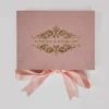 A top-down flat lay of a dusty rose wedding invitation suite on a grey background. The set includes a rose velvet rectangular box with a gold-foiled royal crest and the names tied with a matching pink satin bow.