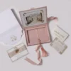 A high-angle view of an open dusty rose velvet wedding invitation box and its coordinating stationery. The inside lid of the box displays a vintage-style illustration of a grand estate framed with gold flourishes. The velvet-lined interior has a recessed compartment and a pink satin ribbon with a matching silk tassel.