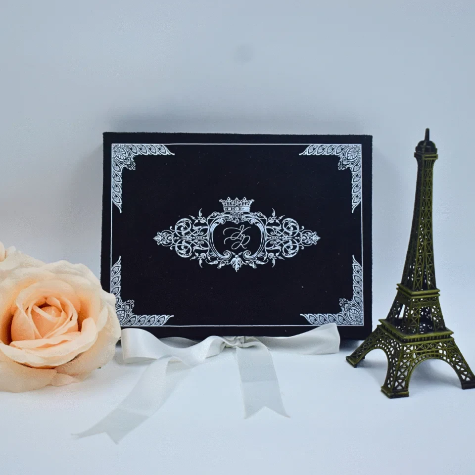 A front-facing view of a rectangular, velvet wedding invitation box set against a light blue background. The lid is decorated with a shimmering silver foil crest featuring a crown and the initials in the center, complemented by intricate silver scrollwork in each of the four corners. The box is elegantly styled with: A white satin ribbon tied in a bow at the base. A small, dark bronze Eiffel Tower figurine standing to the right. A large, soft peach rose partially visible on the left side of the frame.