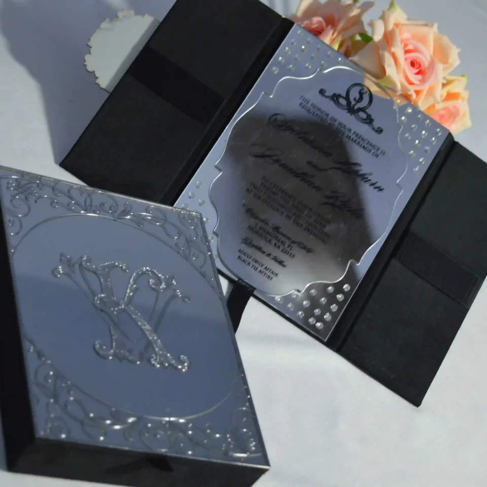 A high-angle shot of an open, champagne-colored silk wedding invitation Box featuring a large, ornate gold crystal-embellished brooch on the left panel. The suite includes: A clear acrylic wedding invitation with white script lettering for the name resting inside the folio. A coordinating ivory envelope with a gold-foiled return address on the flap.The arrangement is decorated with a peach-colored rose in the upper-right corner and a vintage gold lantern in the upper-left, all set against a clean white surface.