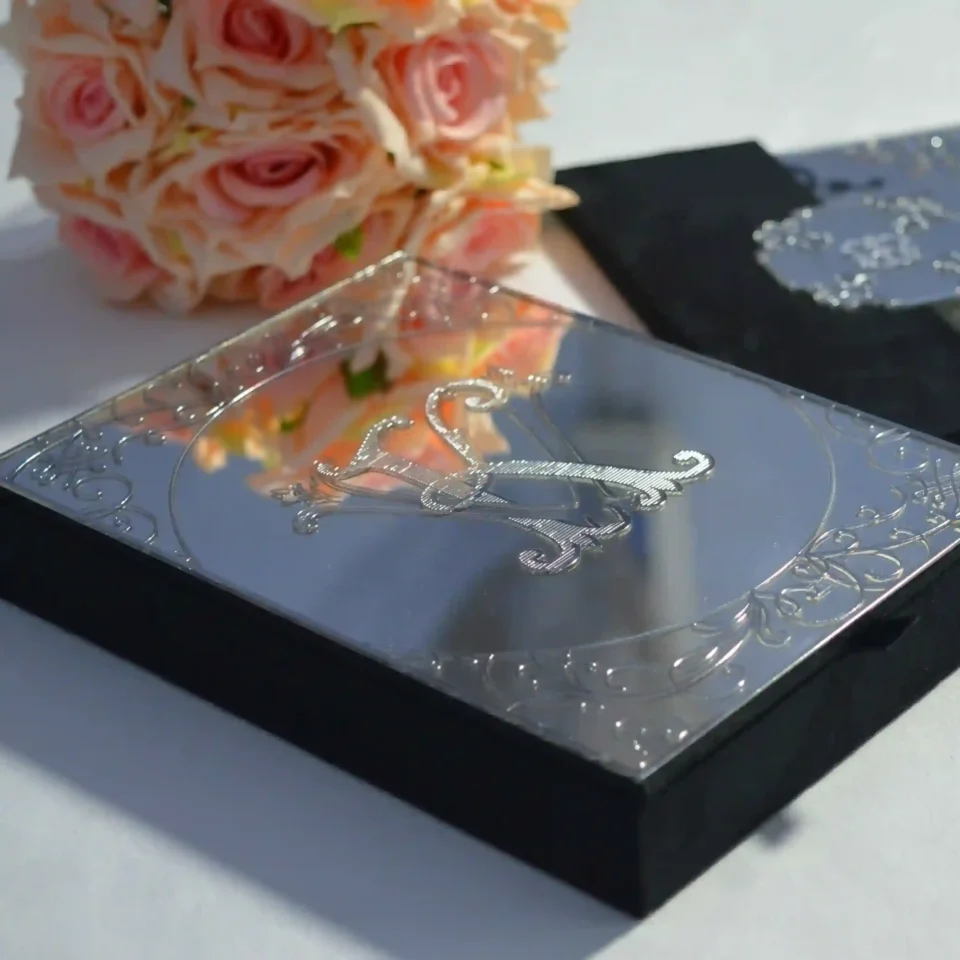 A high-angle shot of an open, champagne-colored silk wedding invitation Box featuring a large, ornate gold crystal-embellished brooch on the left panel. The suite includes: A clear acrylic wedding invitation with white script lettering for the name resting inside the folio. A coordinating ivory envelope with a gold-foiled return address on the flap.The arrangement is decorated with a peach-colored rose in the upper-right corner and a vintage gold lantern in the upper-left, all set against a clean white surface.