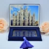 A high-angle view of a luxurious, velvet wedding invitation box shown both closed and open against a light blue background. The set includes:Closed Box:Features a large, shimmering gold foil crest on the lid with the letter in the center, surrounded by intricate floral scrollwork. Open Box:The inner lid displays a gold-etched botanical illustration of a garden with palm trees and a glass conservatory. A matching satin ribbon is used as a pull-tab for the recessed compartment.Stationery: A clear acrylic invitation with white script text leans against a soft peach rose on the left.A small, bronze-colored Eiffel Tower figurine stands to the right, adding a travel-themed touch to the presentation.