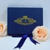 A high-angle view of a luxurious, velvet wedding invitation box shown both closed and open against a light blue background. The set includes:Closed Box:Features a large, shimmering gold foil crest on the lid with the letter in the center, surrounded by intricate floral scrollwork. Open Box:The inner lid displays a gold-etched botanical illustration of a garden with palm trees and a glass conservatory. A matching satin ribbon is used as a pull-tab for the recessed compartment.Stationery: A clear acrylic invitation with white script text leans against a soft peach rose on the left.A small, bronze-colored Eiffel Tower figurine stands to the right, adding a travel-themed touch to the presentation.