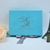 A front-facing view of a rectangular, light turquoise velvet wedding invitation box set against a white background. The lid is personalized with the couple's initials, in a large, elegant silver script. Below the initials, the name and the wedding date printed in clean silver lettering. The box is styled with:A silver satin ribbon tied in a bow at the base. A small, dark bronze Eiffel Tower figurine standing to the right. A soft **peach-colored rose partially visible on the left side of the frame.