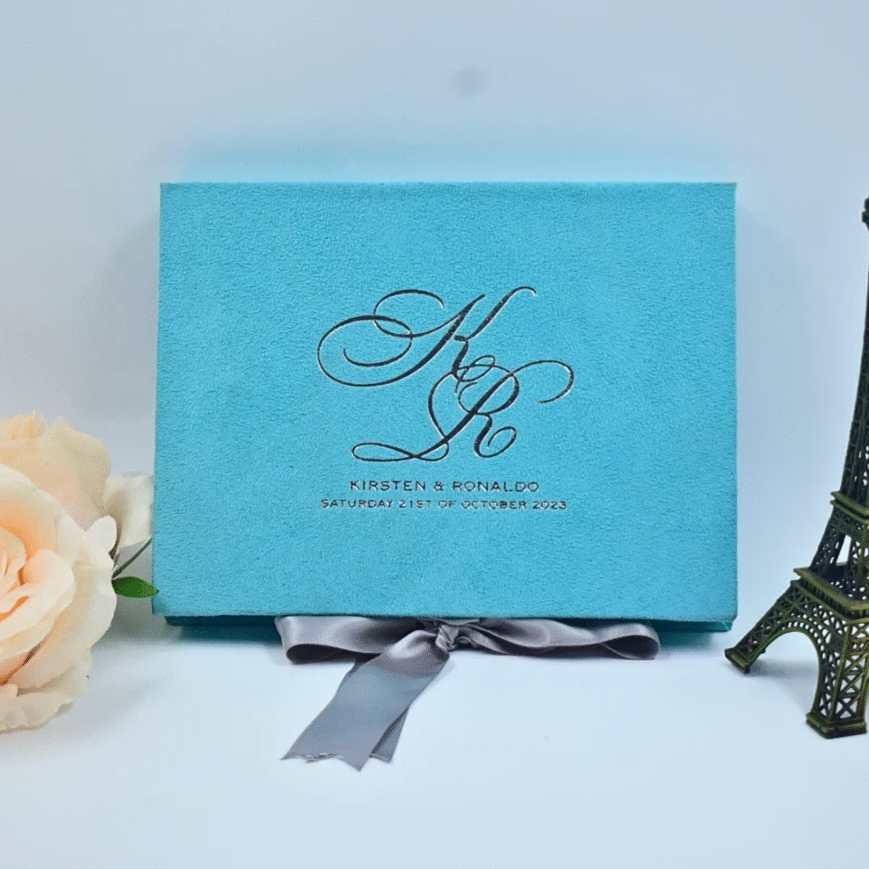 A front-facing view of a rectangular, light turquoise velvet wedding invitation box set against a white background. The lid is personalized with the couple's initials, in a large, elegant silver script. Below the initials, the name and the wedding date printed in clean silver lettering. The box is styled with:A silver satin ribbon tied in a bow at the base. A small, dark bronze Eiffel Tower figurine standing to the right. A soft **peach-colored rose partially visible on the left side of the frame.
