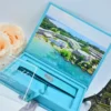 An open light turquoise velvet wedding invitation box shown against a white background. The interior lid features a full-color, panoramic aerial photograph of a tropical beach resort with white buildings, blue swimming pools, and crystal-clear turquoise water. The suite includes: A clear acrylic invitation leaning against a peach rose on the left, featuring white script and serif typography. A recessed compartment in the base of the box, containing a matching turquoise fabric band secured with a silver wax seal. * A light grey satin ribbon used as a pull-tab and decorative element at the base. A small,dark bronze Eiffel Tower figurine standing to the right of the box.