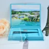An open light turquoise velvet wedding invitation box shown against a white background. The interior lid features a full-color, panoramic aerial photograph of a tropical beach resort with white buildings, blue swimming pools, and crystal-clear turquoise water. The suite includes: A clear acrylic invitation leaning against a peach rose on the left, featuring white script and serif typography. A recessed compartment in the base of the box, containing a matching turquoise fabric band secured with a silver wax seal. * A light grey satin ribbon used as a pull-tab and decorative element at the base. A small,dark bronze Eiffel Tower figurine standing to the right of the box.