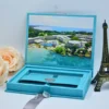 An open light turquoise velvet wedding invitation box shown against a white background. The interior lid features a full-color, panoramic aerial photograph of a tropical beach resort with white buildings, blue swimming pools, and crystal-clear turquoise water. The suite includes: A clear acrylic invitation leaning against a peach rose on the left, featuring white script and serif typography. A recessed compartment in the base of the box, containing a matching turquoise fabric band secured with a silver wax seal. * A light grey satin ribbon used as a pull-tab and decorative element at the base. A small,dark bronze Eiffel Tower figurine standing to the right of the box.