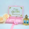 An open blush pink velvet invitation box displayed against a clean white background, part of a "Sweet Sixteen" celebration suite. The presentation includes: * **Inside Lid:** A white card with a decorative gold-glittered border. It features the inspirational quote, She leaves a little Sparkle wherever she goes,printed in elegant gold script and serif fonts. The Base: A recessed compartment holding a clear acrylic invitation with gold-etched details for Samantha Grace's Sweet Sixteen." The invitation is accented with a delicate pink silk tassel. Decorative Accents: A soft peach-colored rose is positioned on the left, and a small golden lantern with blue glass panes stands to the right. A pink satin ribbon pull-tab extends from the front of the box. The combination of soft velvet, gold glitter, and acrylic elements creates a youthful yet sophisticated and luxurious aesthetic.